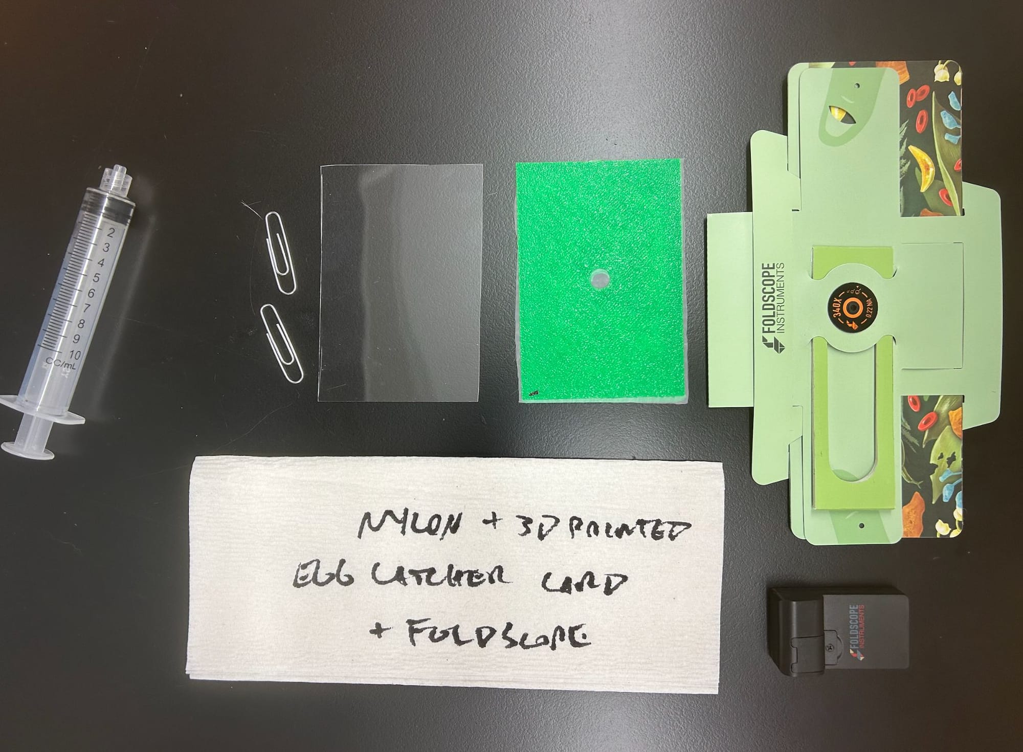 A flat lay of a syringe, paperclips, an acrylic cover, a green nylon egg-catcher card, a Foldscope, and a napkin labeled nylon plus 3D printed egg catcher card plus Foldscope