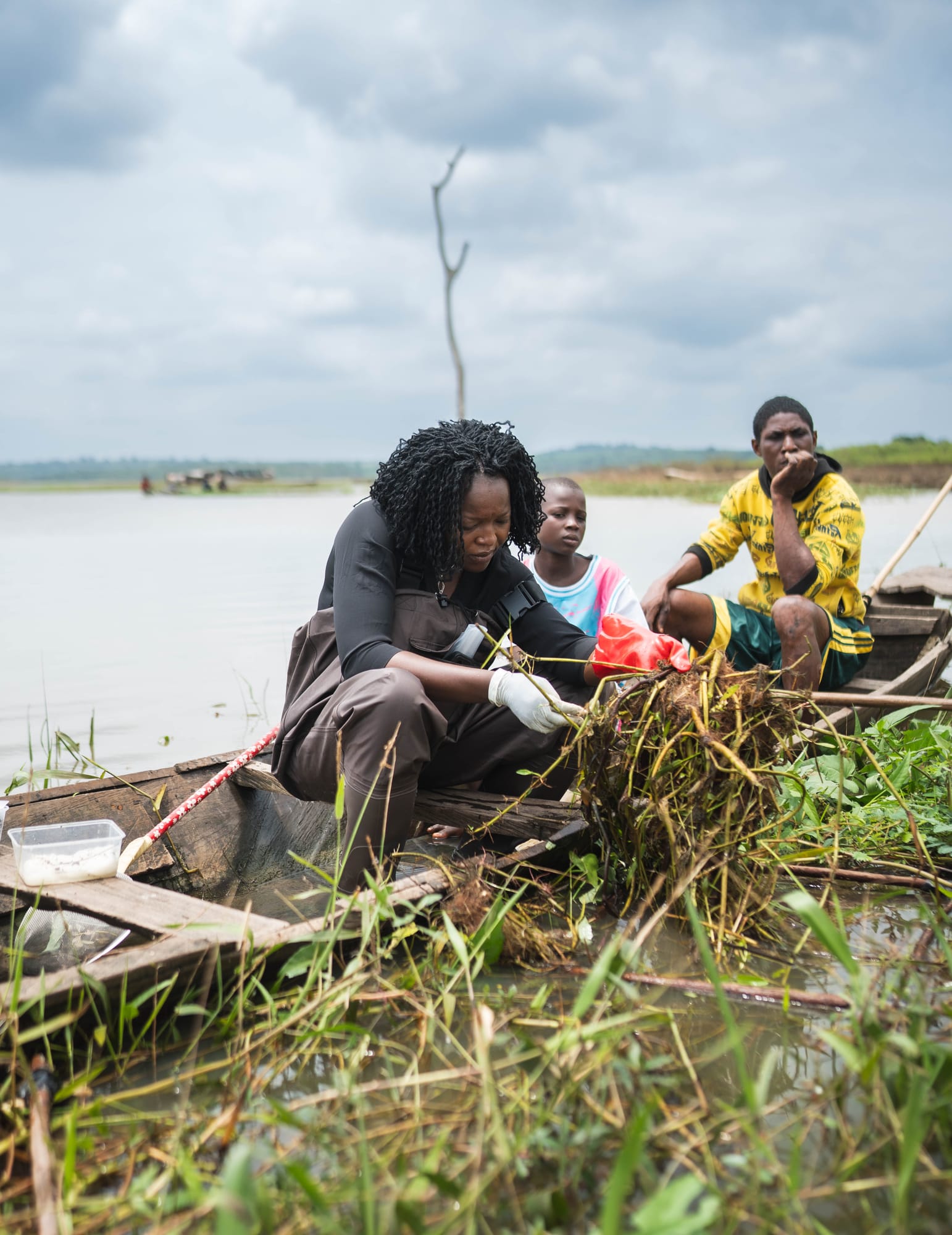 Dr. Olubukola Adelakun kneels in a wooden boat, sorting through a mat of aquatic plants pulled from the reservoir