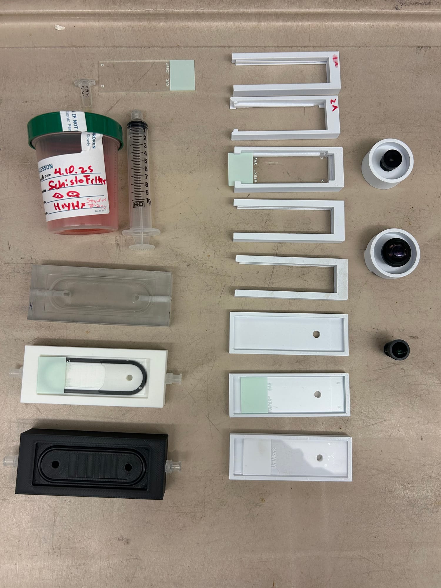 Flat lay of prototypes: 3D-printed slide holders and filter bodies, glass slides, fish-eye lenses, and a specimen cup
